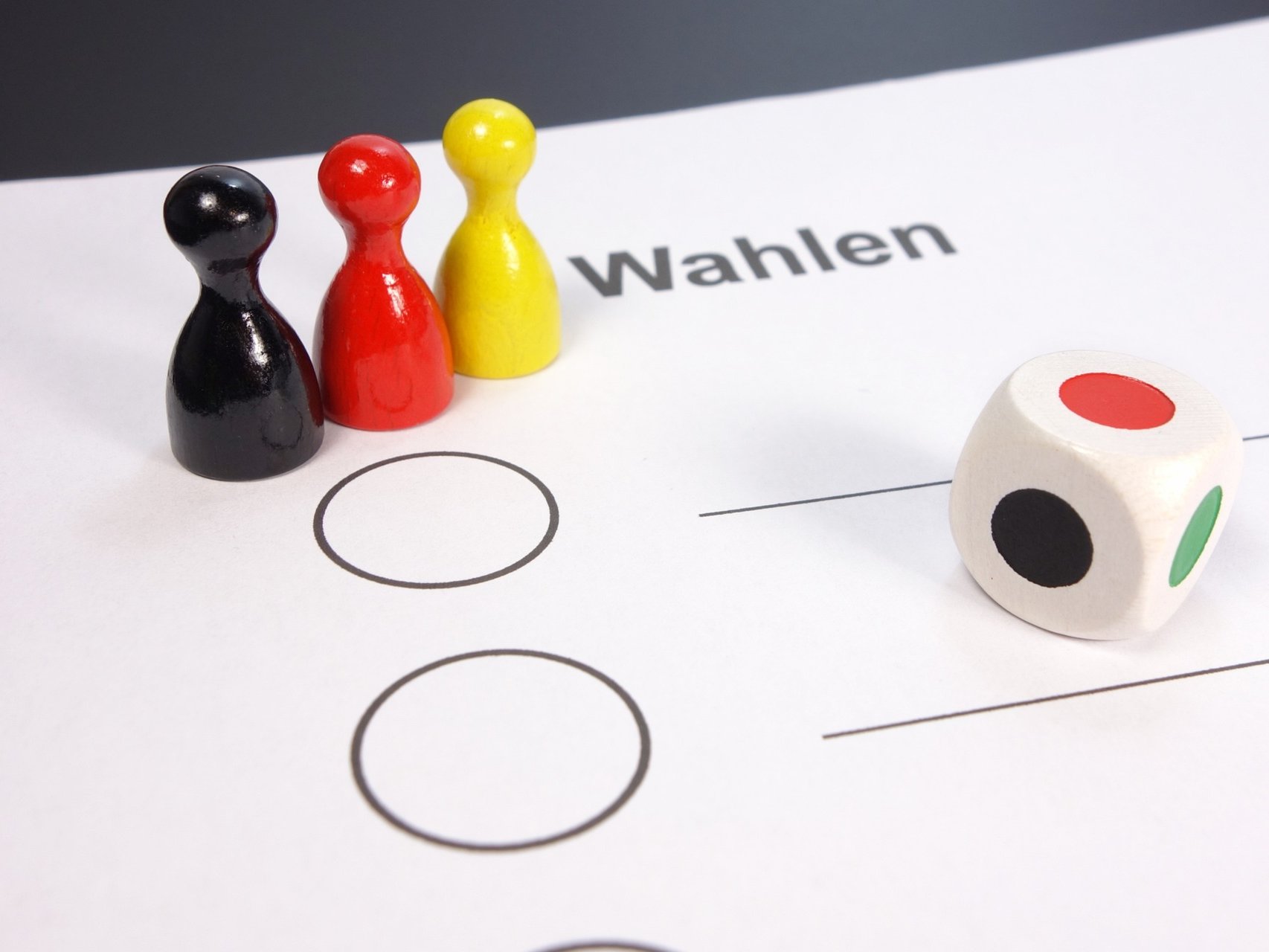 Schwarz rot gelbe Spielfiguren stehen auf einem Wahlzettel im Vordergrund ist ein Farbwürfel zu sehen.