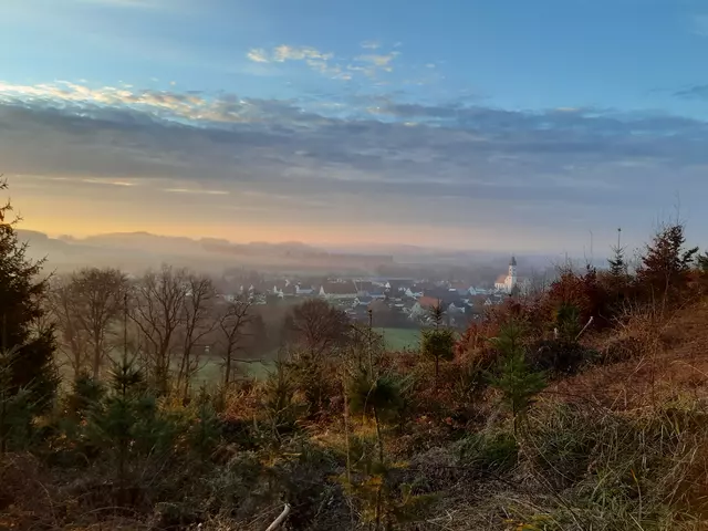 Blick vom Berg über einen herbstlichen Wald auf einen im Nebel liegenden Ort, darüber ist ein blauer Himmel mit einem Streifen Sonnenlicht zu sehen.