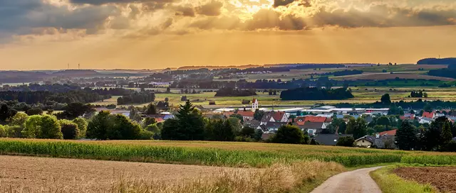 Blick vom Busenberg ins Tal auf die Gemeinde Hochdorf, im Vordergrund liegen grüne Wiesen und goldene Felder, das Dorf in der Mitte des Bildes wird von einer goldgelben Abendsonne, die durch Wolken bricht beschienen.